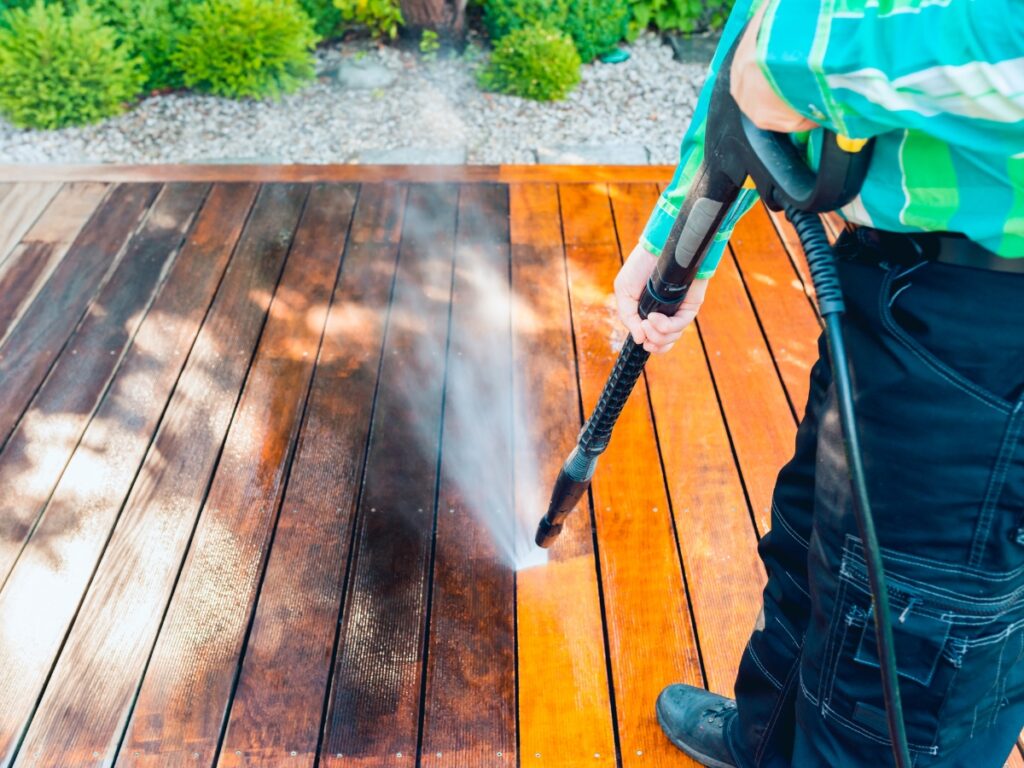 Person using a pressure washer for effective algae and moss removal on a wooden deck, with a clear contrast between cleaned and uncleaned sections.