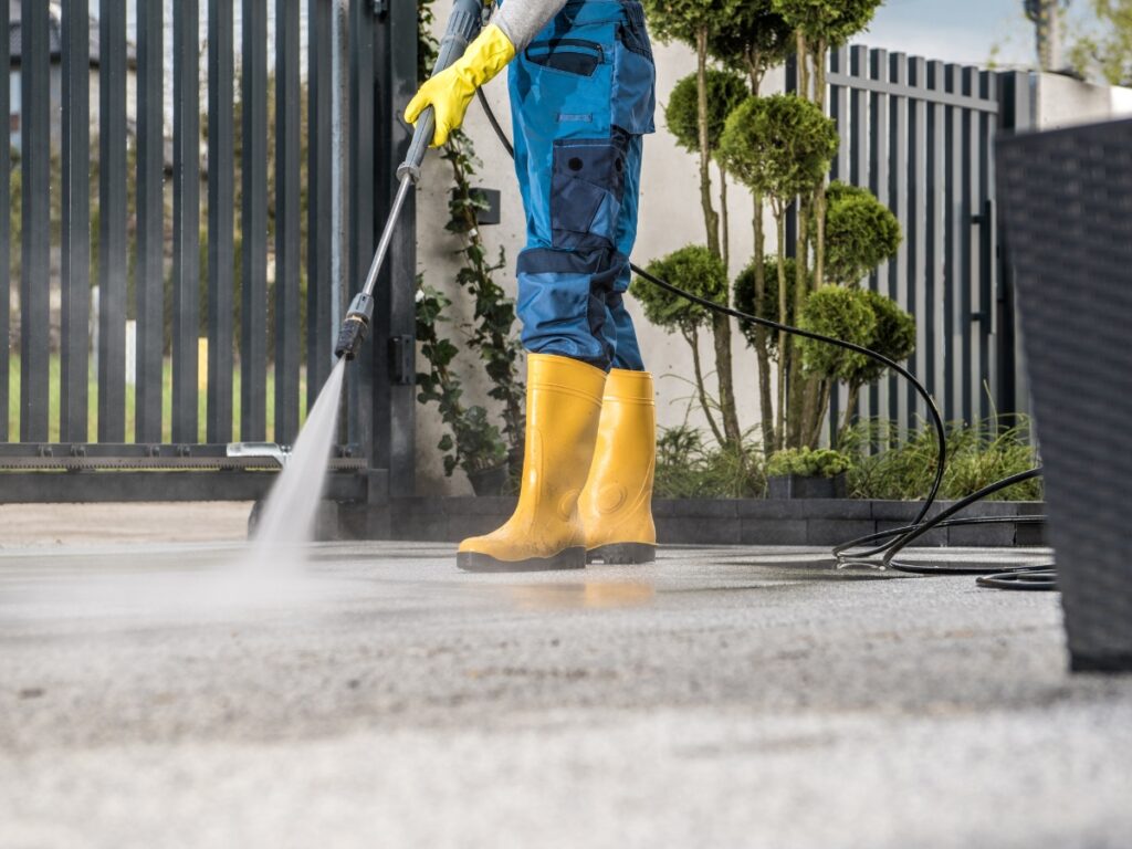 Person wearing yellow boots and gloves uses a pressure washer for algae moss removal, cleaning an outdoor surface near a metal gate and trimmed bushes.