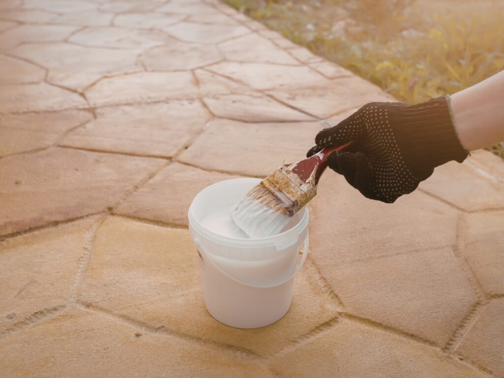 A gloved hand dips a paintbrush into a white plastic bucket filled with white paint, preparing for sealing hardscapes on a textured stone surface outdoors.