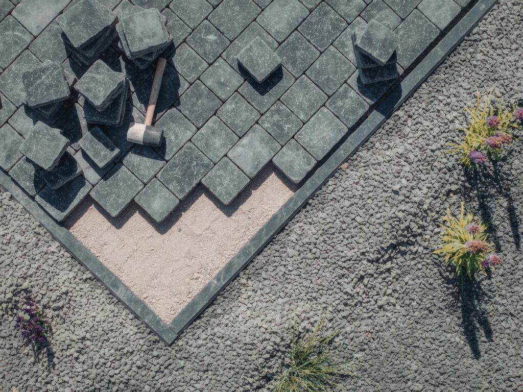 Gray paving stones are being installed in a geometric pattern on a gravel surface, with a rubber mallet and some plants nearby, creating an ideal foundation for sealing hardscapes to enhance durability and appearance.