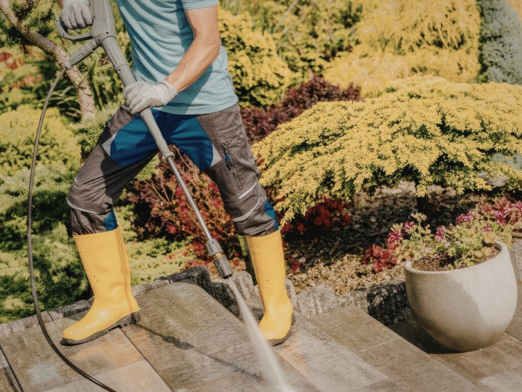 Person in yellow boots uses a pressure washer to clean outdoor steps in a garden, with yellow foliage and a potted plant visible—an essential step before sealing hardscapes for lasting protection.