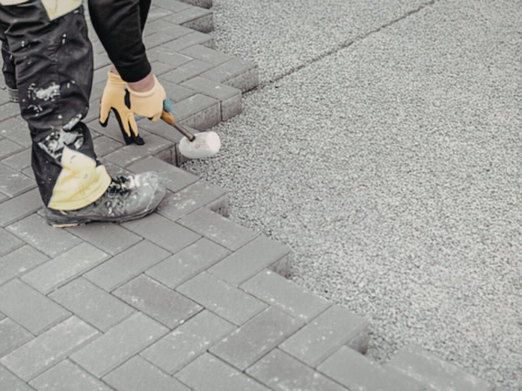 Person wearing work gloves and boots uses a rubber mallet to install gray pavement bricks on a gravel base outdoors, preparing the surface for sealing hardscapes.