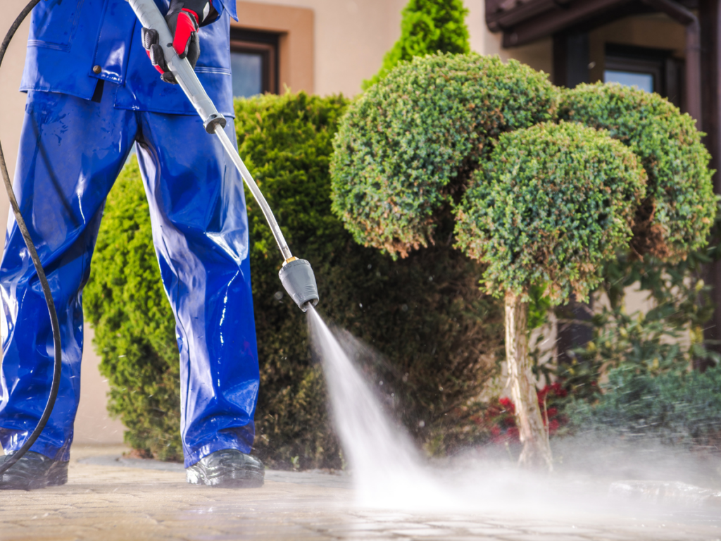 Person in blue waterproof clothing using a pressure washer for cleaning and weed control on an outdoor stone surface near green, manicured bushes.