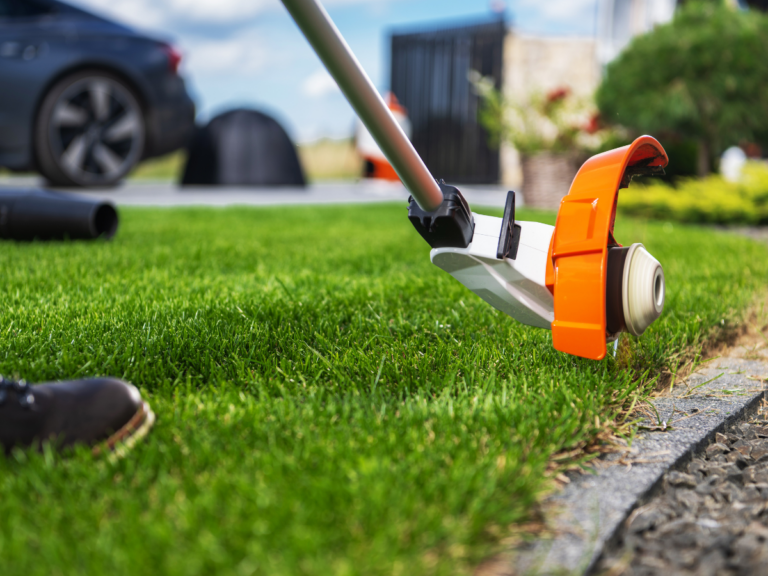 A person uses a string trimmer for weed control to edge a neatly cut lawn next to a stone border in a residential yard.