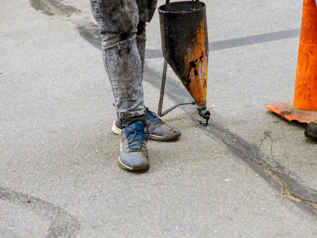 Person performing crack repair, sealing a pavement crack with a hot tar applicator; only their legs and tool are visible next to an orange traffic cone.