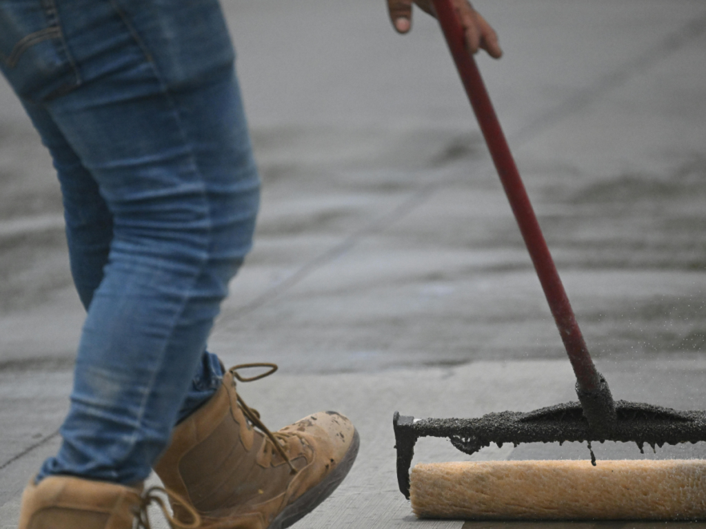 A person wearing jeans and work boots uses a long-handled roller to apply a crack repair substance to a concrete surface.