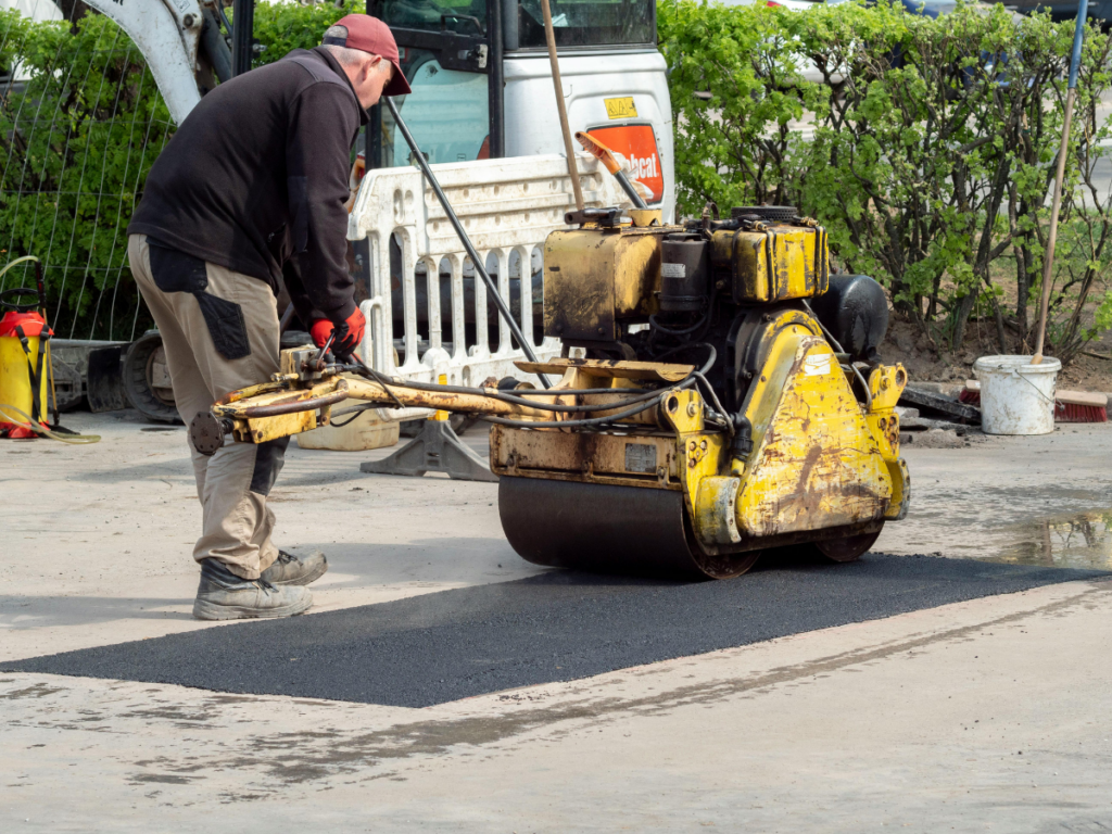 A worker operates a yellow road roller to compact fresh asphalt on a section of pavement at a construction site, following recent crack repair.