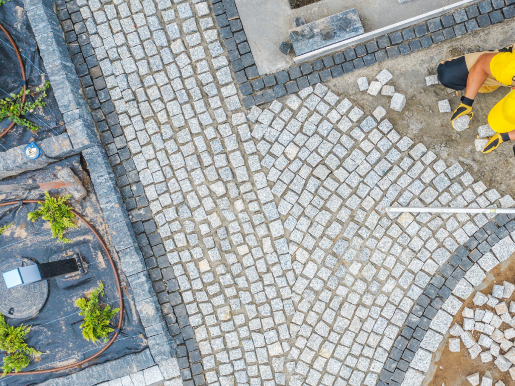 Aerial view of workers in yellow helmets laying patterned stone pavers for a walkway, with plants, construction materials, and crack repair underway nearby.