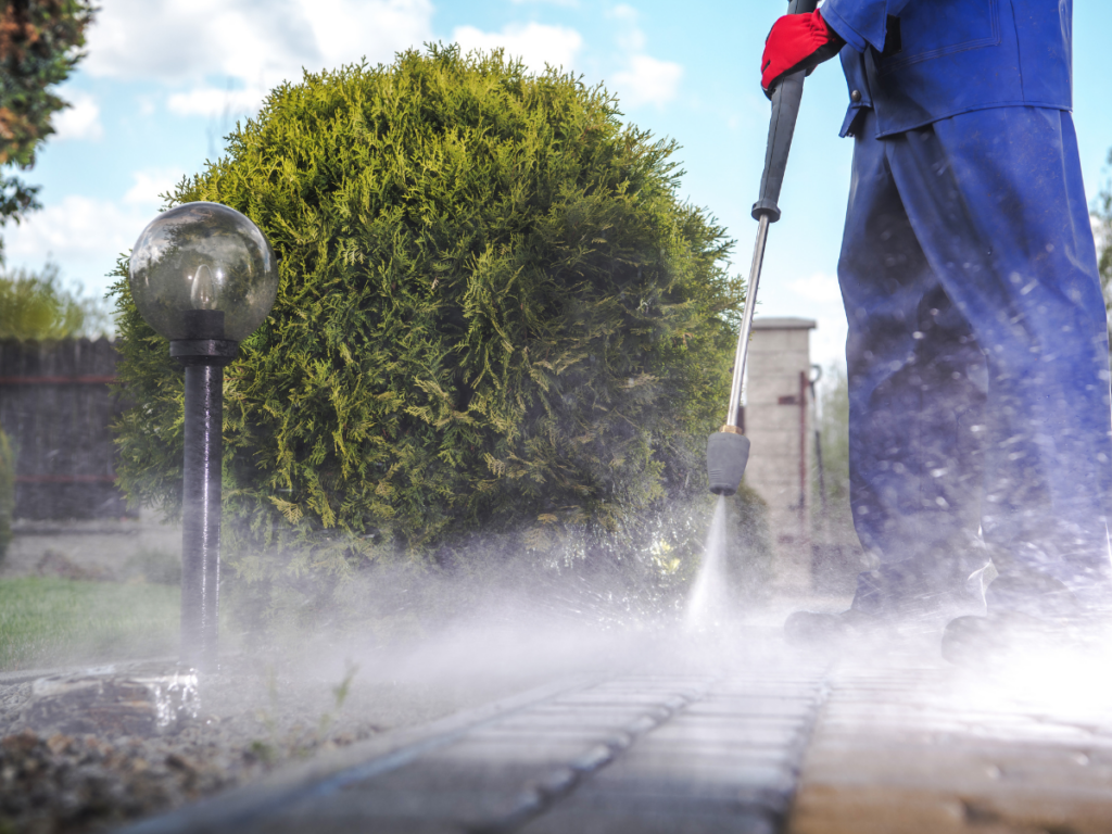 A person in blue clothing and red gloves is pressure washing an outdoor walkway beside a bush and a garden lamp.
