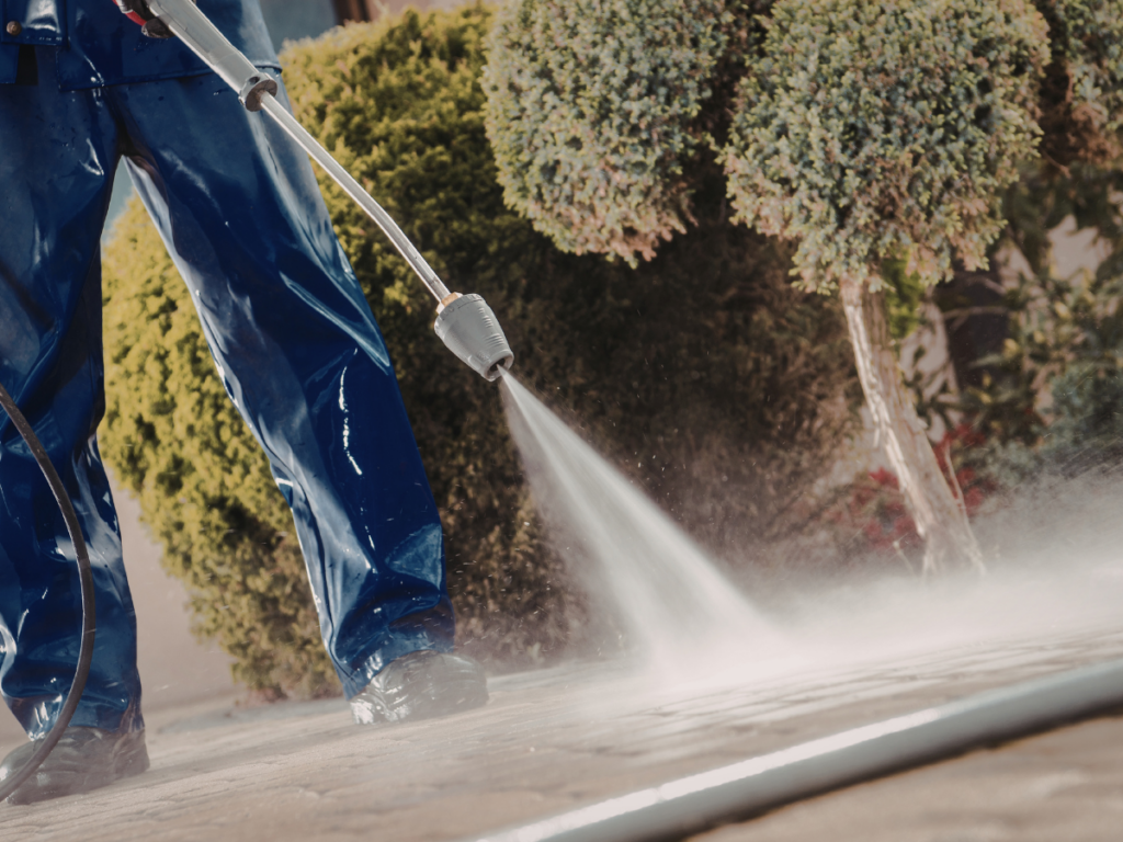 Person in blue waterproof clothing using a pressure washing machine to clean an outdoor pavement, with green bushes in the background.