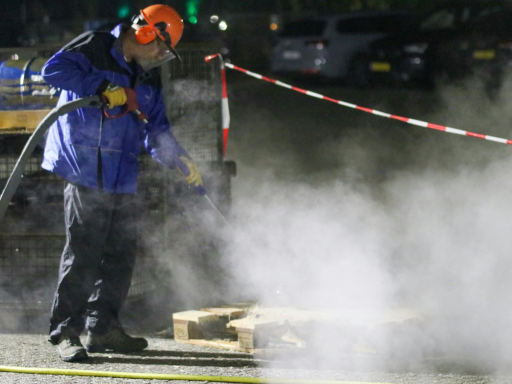 A person in safety gear is pressure washing wooden pallets outdoors at night, with steam rising and caution tape visible in the background.