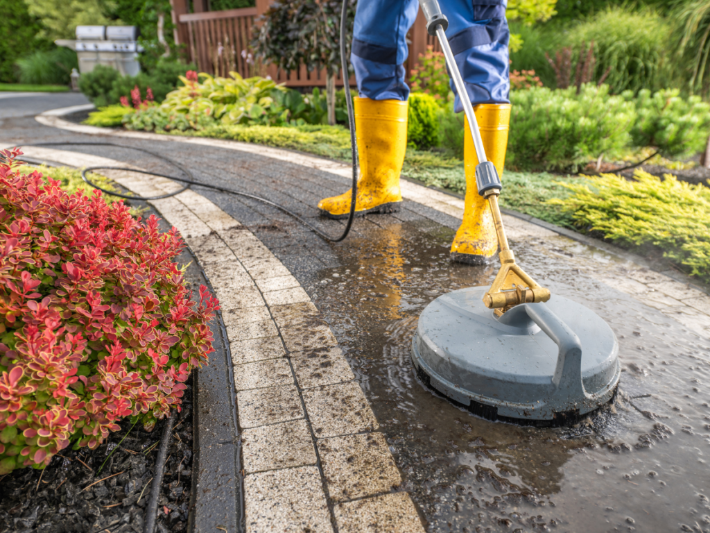 Wearing yellow boots, a person uses a pressure washing surface cleaner to tackle a stone garden pathway surrounded by lush plants and shrubs.