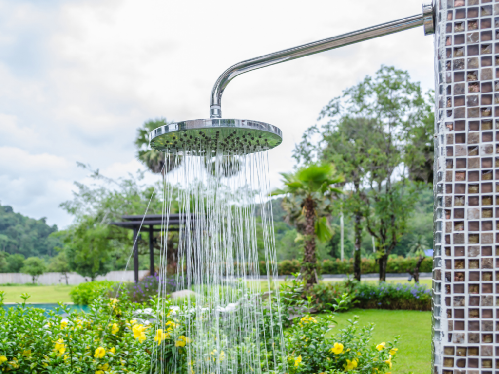 An inviting outdoor shower with water running is mounted on a tiled wall, surrounded by lush green plants, vibrant yellow flowers, and trees in a peaceful garden setting.