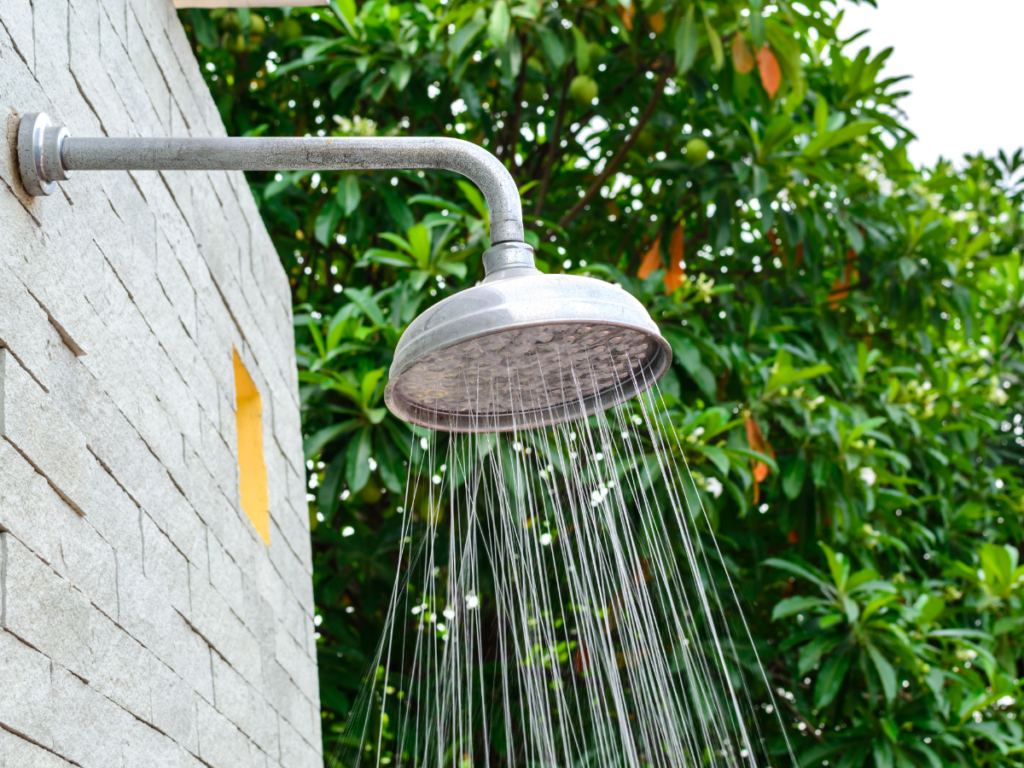 An inviting outdoor showerhead attached to a stone wall, spraying water, with green leafy trees in the background creates a refreshing outdoor shower experience nestled in nature.