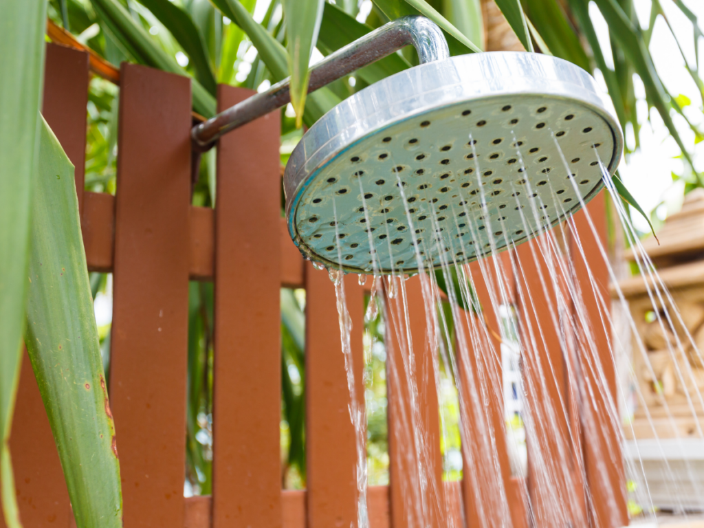Outdoor showerhead with water flowing, mounted on a metal pipe near a wooden fence and green plants in the background, creating an inviting outdoor shower experience.