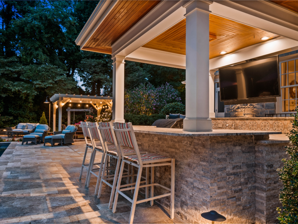 Outdoor patio with a stone bar, four barstools, a mounted TV, and a covered seating area with string lights—this inviting space embodies the charm of outdoor bars. Surrounded by trees in the evening, it’s perfect for relaxing or entertaining outdoors.