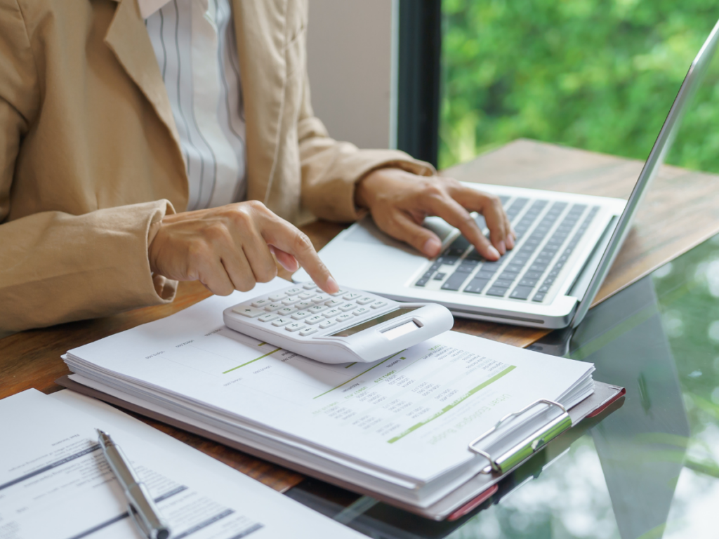 Person in business attire uses a calculator and laptop at a desk, with financial documents and reports on a clipboard. Through the window in the background, lush green foliage and hints of nearby outdoor bars create a lively yet professional atmosphere.