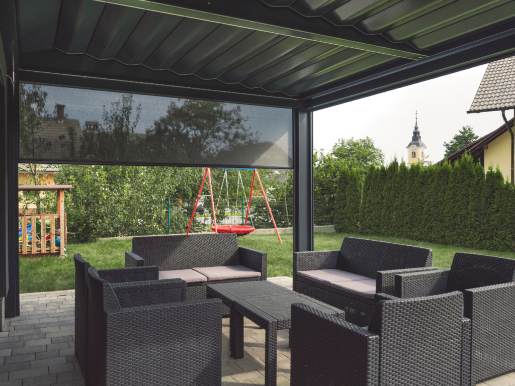 Outdoor seating area with black wicker furniture under stylish patio covers, facing a grassy yard with a children’s swing set and a wooden playhouse, surrounded by hedges and houses.