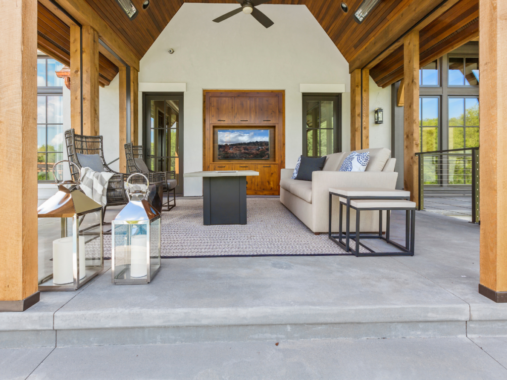 Open-air patio with wooden beams and stylish patio covers, a beige sofa, black chairs, two lanterns, and a wall-mounted TV facing a coffee table on a large rug. Floor-to-ceiling windows provide outdoor views.