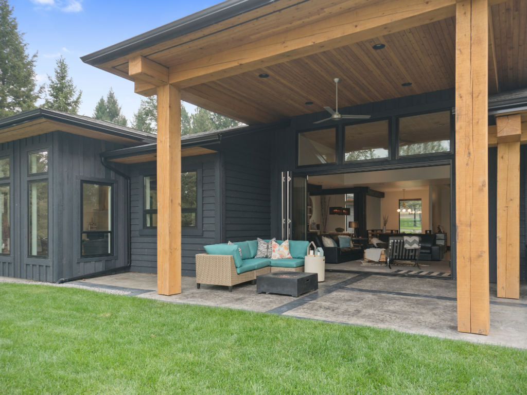Modern house with dark exterior, wooden beams, patio covers for shade, outdoor seating, large sliding doors, and view into living area, surrounded by green lawn and trees.