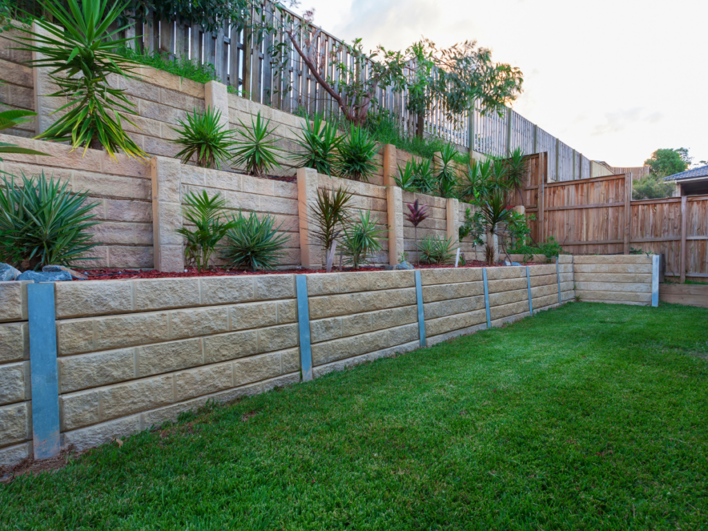 Tiered retaining walls with stone blocks and metal supports, landscaped with green plants and shrubs, bordered by a wooden fence and a well-maintained grass lawn, offering both beauty and easy retaining wall maintenance for lasting curb appeal.