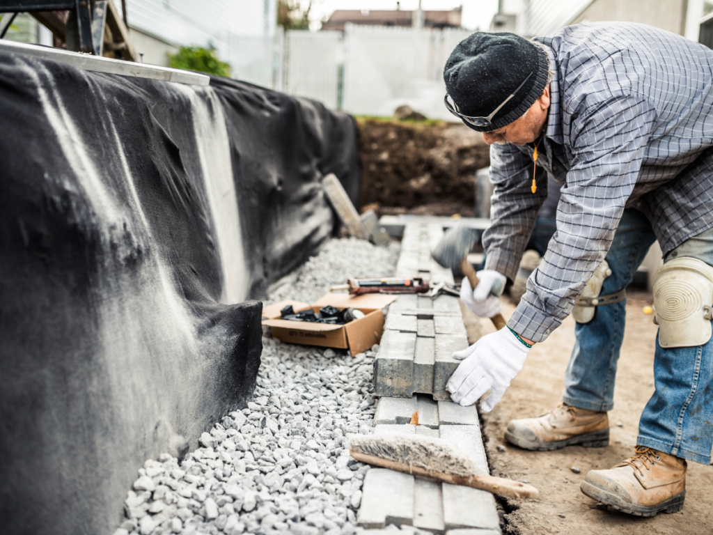 A person wearing gloves and a beanie is laying bricks and tapping them into place along a gravel path at a construction site, carefully focusing on retaining wall maintenance.