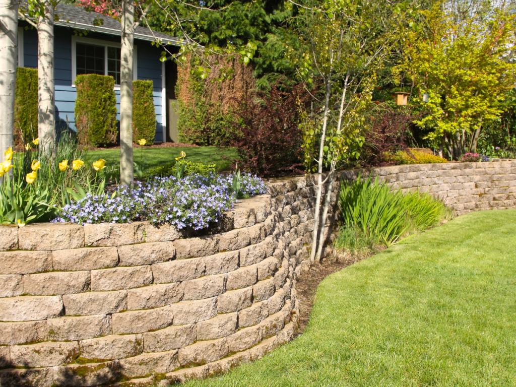 A landscaped yard with curved retaining walls made of stacked stone blocks showcases meticulous retaining wall maintenance, complemented by flower beds, green shrubs, trees, and a well-maintained lawn in front of a house.