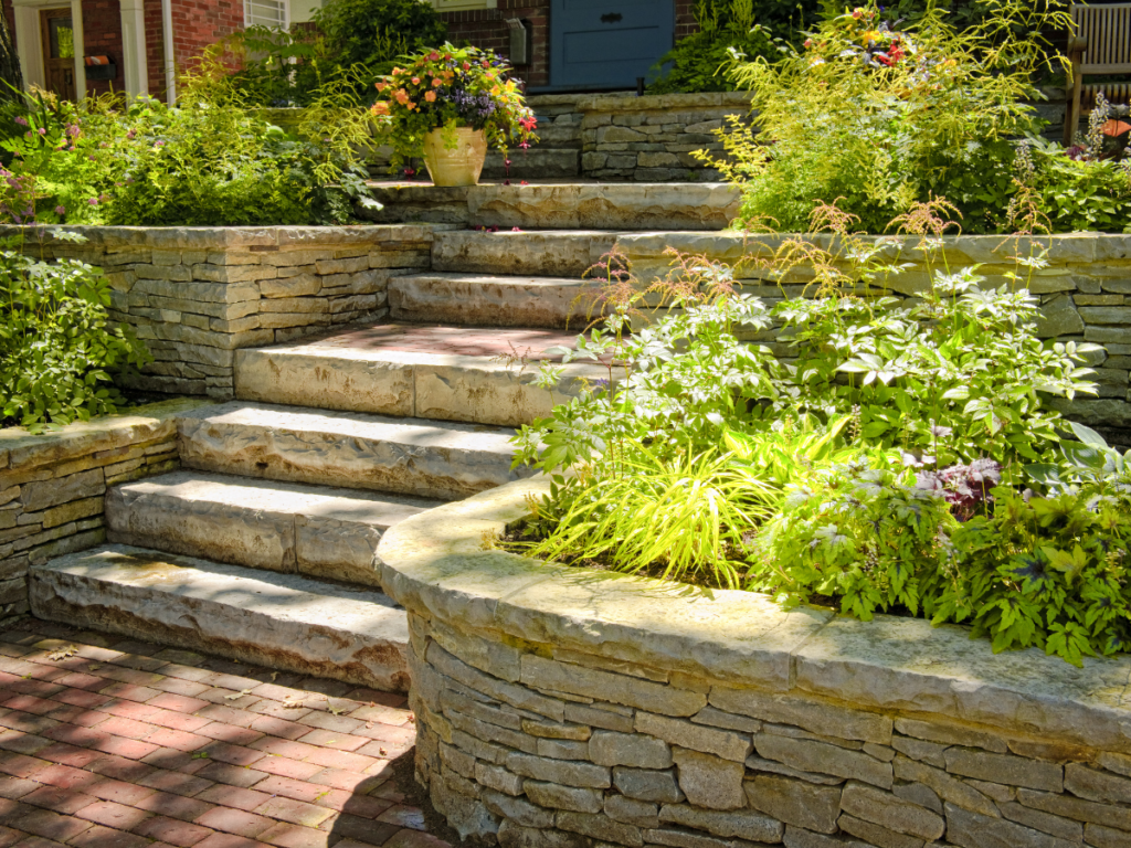 Stone steps lead up through terraced garden beds with green plants and colorful flowers, bordered by well-kept stacked stone walls that highlight careful retaining wall maintenance, on a sunny day.